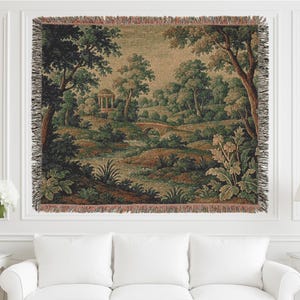Op de afbeelding: Een geweven wandtapijt met een serene landschap met bomen, een brug en een kleine structuur. Het wandtapijt heeft een kleurenpalet van groen, bruin en beige, met een gefranjerde rand. Het kunstwerk is ingelijst en wordt boven een witte bank getoond.