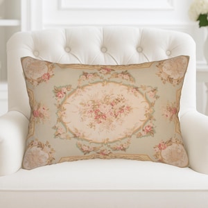Pode incluir: Almofada retangular decorativa com um padrão floral. A almofada apresenta um design oval central com rosas cor-de-rosa e detalhes verdes sobre um fundo verde claro. Está colocada numa poltrona branca.