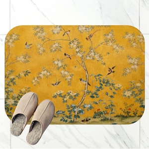 May include: A rectangular bath mat with a mustard yellow background featuring a detailed print of flowering branches, green foliage, and various birds in flight and perched. A pair of light beige slip-on slippers with grey interiors are placed on top.