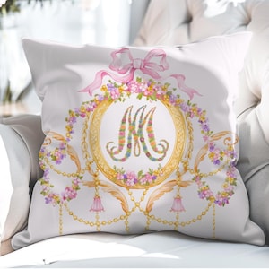 May include: A white square throw pillow featuring a central gold circular frame containing a floral letter M. The design includes a pink ribbon bow at the top, cascading gold chains, and decorative floral garlands in shades of pink, yellow, and purple.