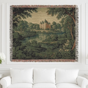 Può includere: Coperta arazzo tessuta raffigurante un paesaggio con un castello, alberi e acqua. La coperta ha un bordo con frange ed è principalmente verde, marrone e beige. L'arazzo è esposto su una parete bianca sopra un divano bianco.