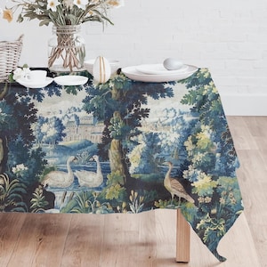 May include: A blue and green tapestry tablecloth with a scenic design featuring swans, trees, and a building. The tablecloth is draped over a wooden table, with a vase of flowers and white plates on top.