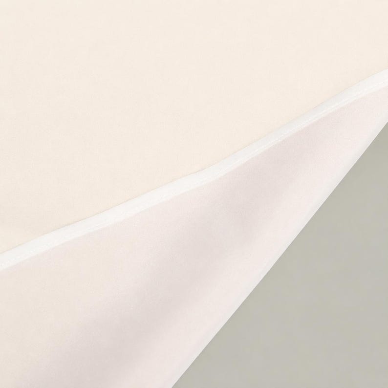May include: A folded piece of cream-colored fabric featuring a smooth, matte texture. The fabric is layered, revealing a clean, straight white stitched hem along the diagonal fold that separates the two sections of the material.