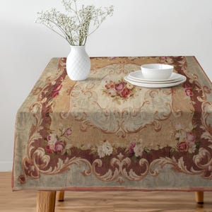 May include: A rectangular tablecloth with a floral design featuring roses in shades of pink and cream, set on a table. The tablecloth has a beige background with a decorative border in shades of brown and burgundy. A white vase with flowers and white plates and a bowl are on the table.