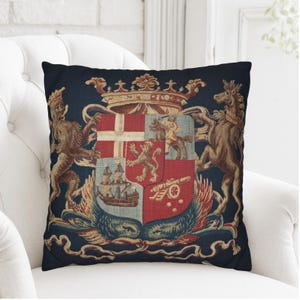 Puede incluir: Un cojín decorativo con un diseño detallado de escudo heráldico. El escudo, con corona, leones, caballos y un barco, se presenta sobre un fondo azul oscuro. El cojín está colocado sobre un sillón blanco.