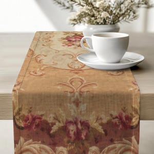 Puede incluir: Un camino de mesa rectangular con fondo beige y un patrón repetitivo de rosas rojo oscuro, hojas de acanto color crema y volutas intrincadas. La tela presenta una apariencia texturizada con un tejido visiblemente marcado.