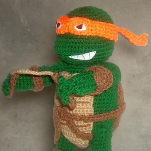 Op de afbeelding: Handgemaakte gehaakte figuur van een Teenage Mutant Ninja Turtle, met een groen lichaam, een oranje masker en bruine accenten. De figuur heeft een lachend gezicht en poseert met uitgestrekte armen. Het ontwerp is geïnspireerd op de populaire tekenfilmserie.