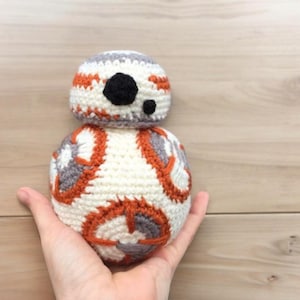 May include: A handmade crochet BB-8 droid toy. The toy is white with orange and gray accents, and a black eye. The head is gray and white. The toy is held in a hand.