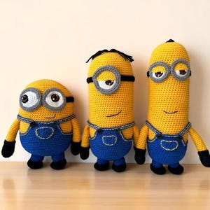 May include: Three handmade crochet Minion dolls. Each Minion is yellow with blue overalls, black shoes, and black gloves. They have large eyes and wear black goggles. The Minions are standing on a wooden surface.