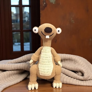 Puede incluir: Un peluche de ganchillo hecho a mano de un personaje de dibujos animados, con un cuerpo marrón, un vientre color crema y detalles blancos. El juguete tiene ojos grandes y redondos, una nariz marrón oscuro y una boca sonriente con dos dientes blancos.