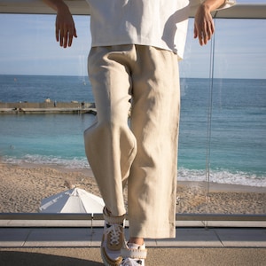 Puede incluir: Una persona con una blusa de lino blanca y pantalones de lino beige, combinados con zapatillas de plataforma. Las zapatillas tienen detalles en blanco, beige y dorado. El conjunto se presenta sobre un fondo de playa y océano.