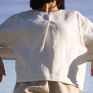 Puede incluir: Una camisa de lino blanca de corte holgado y mangas tres cuartos. La camisa está ligeramente fruncida en el escote.