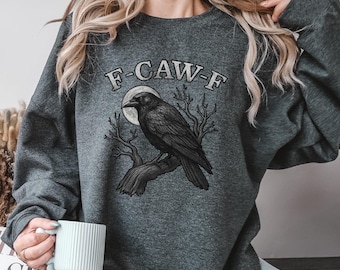 May include: Dark gray sweatshirt featuring a black raven perched on a branch, with a full moon in the background. The text "F-CAW-F" is arched above the raven. The design is printed on a soft, comfortable sweatshirt.