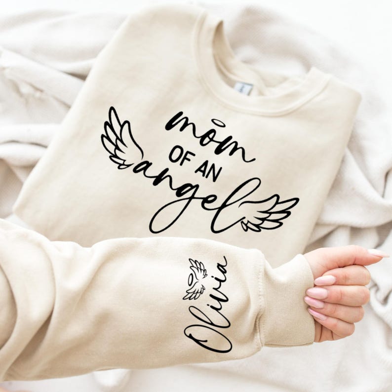 May include: A beige sweatshirt with the text "mom of an angel" and a pair of angel wings on the front. The left sleeve has the name "Olivia" and a small angel wing.