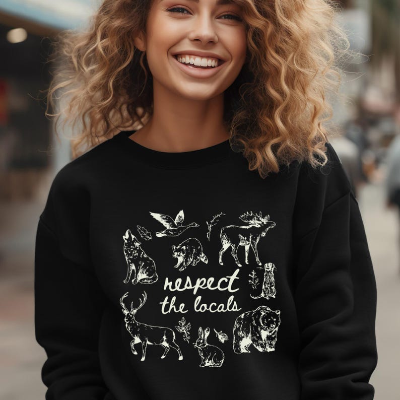 May include: Black sweatshirt featuring a white graphic of various animals, including a wolf, bear, and deer, with the text "respect the locals." The design is a nature-themed illustration.