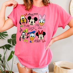 Peut inclure: T-shirt rose avec des visages de personnages de dessins anim&eacute;s, dont Mickey Mouse, Minnie Mouse, Donald Duck et Daisy Duck. Le t-shirt a des manches retrouss&eacute;es et des signatures imprim&eacute;es au-dessus et en dessous des visages.