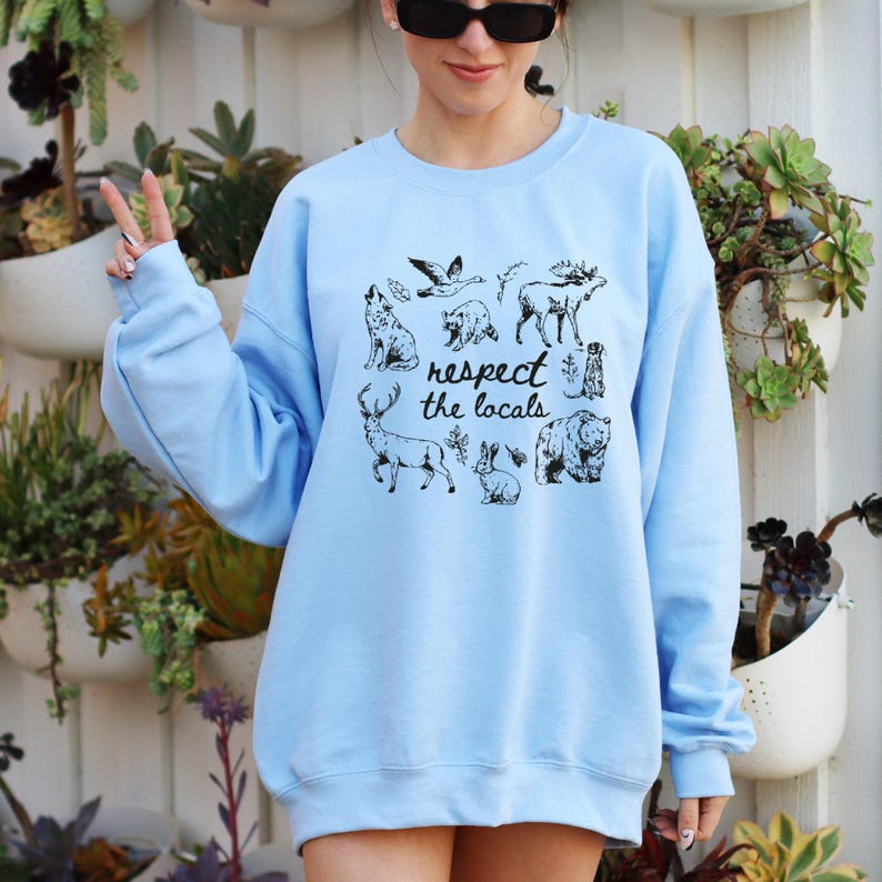May include: Light blue sweatshirt with a black graphic of various animals, including a wolf, bear, and deer. The text "respect the locals" is printed below the animal illustrations. The sweatshirt is a long-sleeved crew neck.