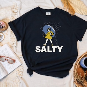 May include: Black t-shirt with a graphic of a person under a blue umbrella in the rain. The person is wearing a yellow dress and the word "SALTY" is printed in white block letters below the graphic.