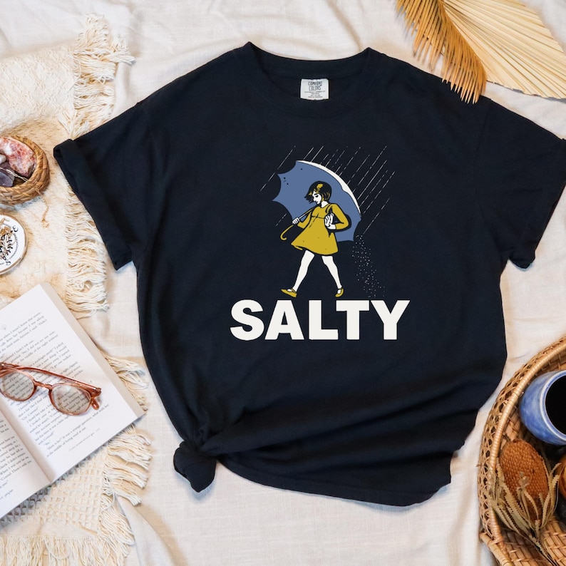 May include: Black t-shirt with a graphic of a person under a blue umbrella in the rain. The person is wearing a yellow dress and the word "SALTY" is printed in white block letters below the graphic.