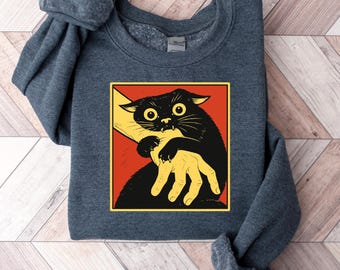 May include: A navy blue sweatshirt featuring a graphic of a black cat biting a yellow hand, set against a red background. The cat has wide eyes and is in a defensive posture. The design is framed in a yellow border.