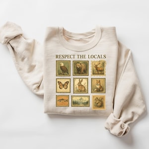 May include: Beige sweatshirt with the text "RESPECT THE LOCALS" above nine postage stamp-style images of wildlife, including an eagle, bear, deer, butterfly, rabbit, fox, fish, mountain scene, and squirrel. The sweatshirt has long sleeves.