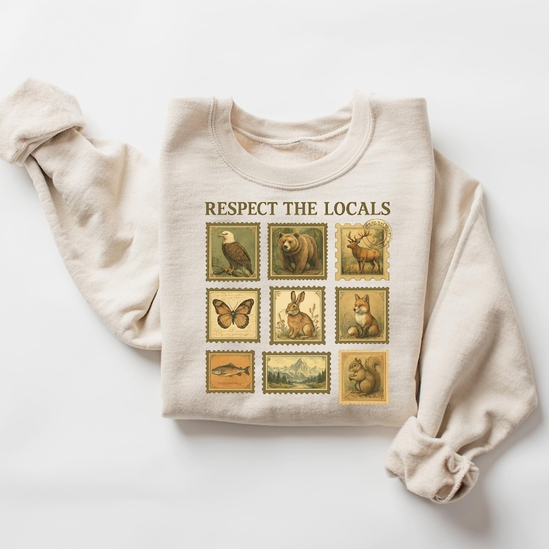 May include: Beige sweatshirt with the text "RESPECT THE LOCALS" above nine postage stamp-style images of wildlife, including an eagle, bear, deer, butterfly, rabbit, fox, fish, mountain scene, and squirrel. The sweatshirt has long sleeves.