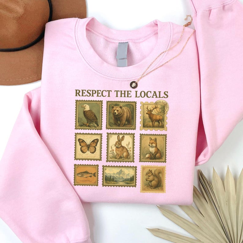 May include: A light pink sweatshirt featuring a design of vintage-style postage stamps with various animals and the text "Respect the Locals". The stamps include an eagle, bear, deer, butterfly, rabbit, fox, fish, mountain scene, and squirrel.
