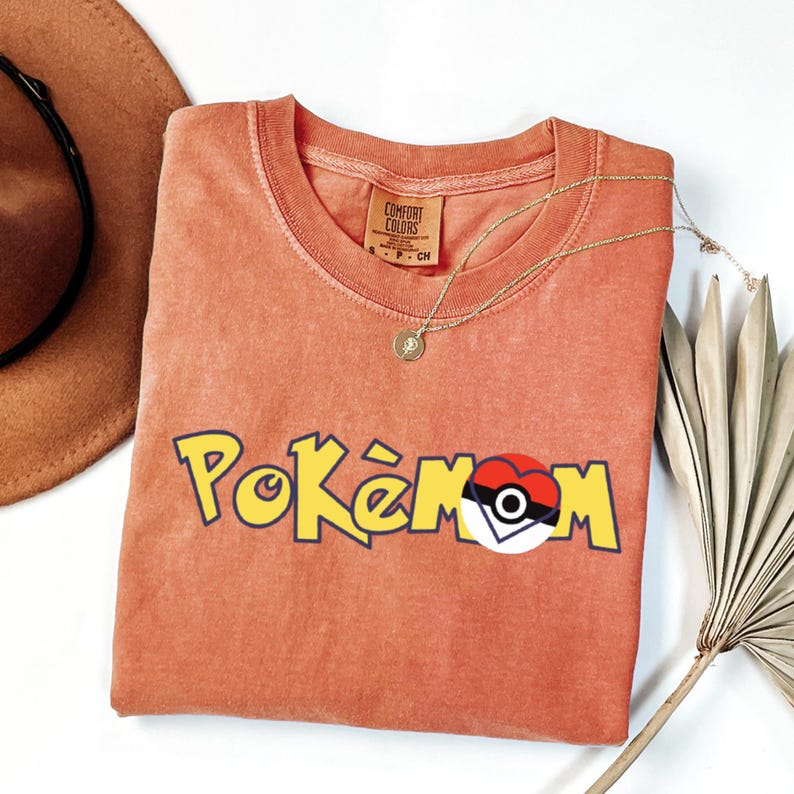 Puede incluir: Una camiseta de color coral con la palabra "Pok&eacute;mon" en letras amarillas con contorno negro. La "O" de Pok&eacute;mon se reemplaza con un dise&ntilde;o de Pok&eacute; Ball. Tambi&eacute;n se ven un collar dorado y un sombrero marr&oacute;n.