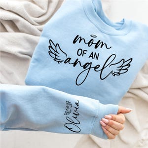 May include: Light blue sweatshirt with the text "mom of an angel" and angel wings printed on the front. The text "Olivia" is printed on the sleeve with a small angel wing.
