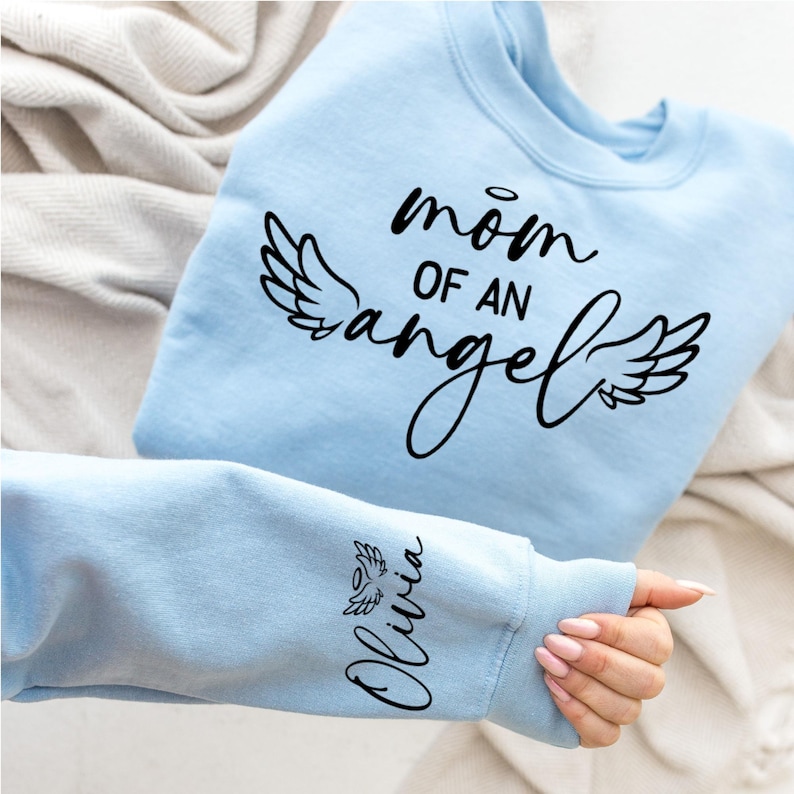 May include: Light blue sweatshirt with the text "mom of an angel" and angel wings printed on the front. The text "Olivia" is printed on the sleeve with a small angel wing.