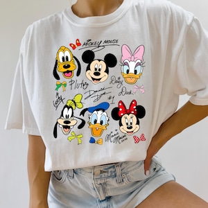 Peut inclure: T-shirt blanc avec des visages de personnages de dessins anim&eacute;s color&eacute;s, dont Mickey Mouse, Minnie Mouse, Donald Duck, Daisy Duck, Pluto et Dingo. Les noms des personnages sont &eacute;crits en cursive.