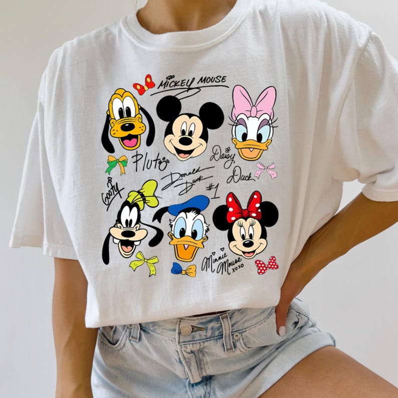 Peut inclure: T-shirt blanc avec des visages de personnages de dessins anim&eacute;s color&eacute;s, dont Mickey Mouse, Minnie Mouse, Donald Duck, Daisy Duck, Pluto et Dingo. Les noms des personnages sont &eacute;crits en cursive.
