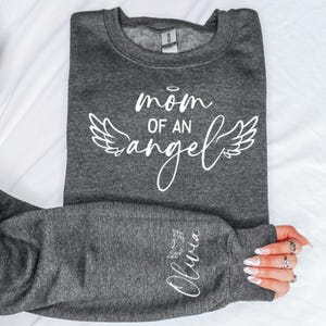 May include: Grey sweatshirt with the text "mom of an angel" in white with angel wings on either side. The left sleeve has the name "Olivia" written in white with a small heart.