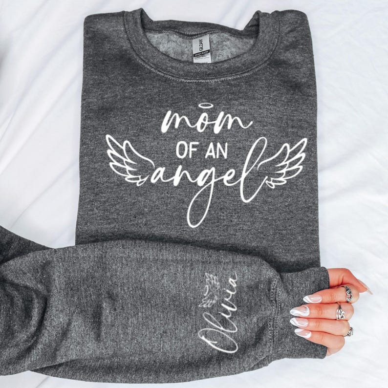 May include: Grey sweatshirt with the text "mom of an angel" in white with angel wings on either side. The left sleeve has the name "Olivia" written in white with a small heart.
