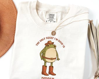 May include: Cream-colored t-shirt featuring a cartoon frog wearing a cowboy hat and boots, with the text "THE ONLY SQUAT I'M DOIN' IS DIDDLY" in brown. A gold necklace and a brown hat are also visible.