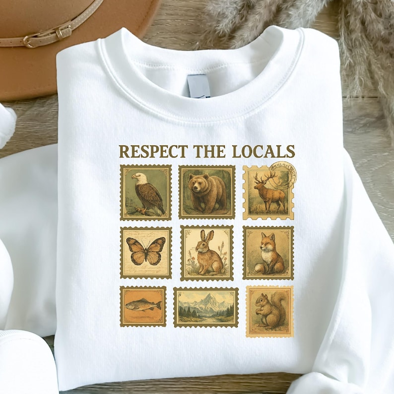 May include: White sweatshirt with the text "RESPECT THE LOCALS" above nine vintage-style postage stamp illustrations of wildlife, including an eagle, bear, deer, butterfly, rabbit, fox, fish, mountains, and squirrel.