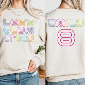 May include: A cream-colored sweatshirt with the text "LET'S GLOW CRAZY" in rainbow-colored block letters on the front. The back of the sweatshirt has the name "EMILY" and the number "8" in a similar rainbow design.