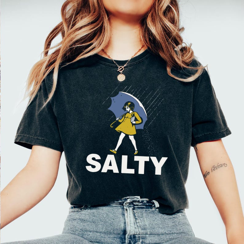 May include: Black t-shirt with the word "SALTY" in white block letters. The shirt features a graphic of a person in a yellow dress and a blue umbrella walking in the rain. The overall design has a vintage, slightly distressed look.