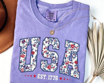 May include: A lavender Comfort Colors t-shirt with the word "USA" in a floral pattern. The shirt also features "EST. 1776" and a gold necklace. A brown hat and dried plant are in the background.