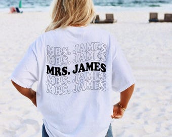 May include: A white t-shirt with the text "MRS. JAMES" in a stacked design on the back. The middle text is in bold black, while the others are outlined in black. The shirt is worn on a beach.