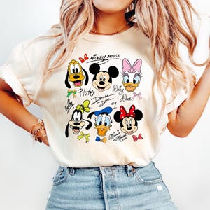 Peut inclure: T-shirt cr&egrave;me avec des personnages Disney : Mickey Mouse, Pluto, Donald Duck, Daisy Duck, Minnie Mouse et Dingo. Le texte "Mickey Mouse" est en haut, avec les noms des personnages en dessous de chaque image.