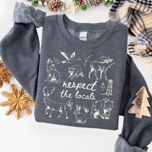 May include: Dark gray sweatshirt with the phrase "respect the locals" and illustrations of various animals, including a wolf, bear, and moose. The sweatshirt has a crew neck and long sleeves.