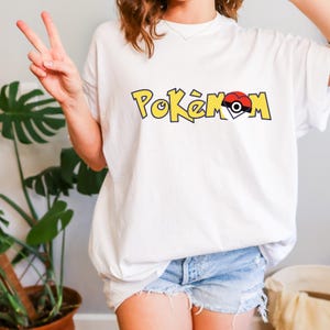 Puede incluir: Camiseta blanca con la palabra "Pok&eacute;mon" en letras amarillas con contorno negro. La "O" se reemplaza con un dise&ntilde;o de Pok&eacute;ball rojo y blanco. La camiseta es de estilo casual, de manga corta.