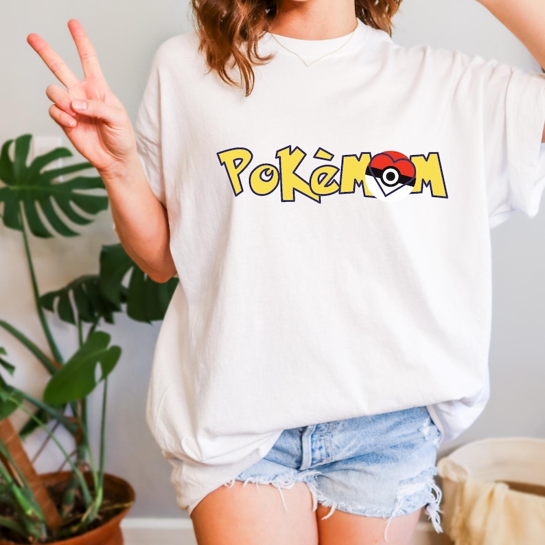 Puede incluir: Camiseta blanca con la palabra "Pok&eacute;mon" en letras amarillas con contorno negro. La "O" se reemplaza con un dise&ntilde;o de Pok&eacute;ball rojo y blanco. La camiseta es de estilo casual, de manga corta.