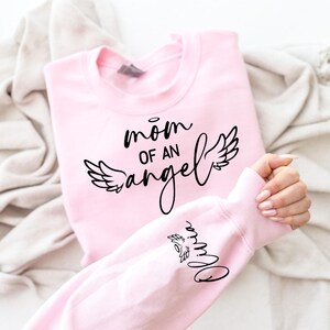 May include: Pink sweatshirt with the text "mom of an angel" and angel wings printed on the front. The text "Olivia" is printed on the sleeve.