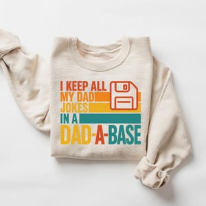 May include: Cream-colored sweatshirt with the text "I KEEP ALL MY DAD JOKES IN A DAD-A-BASE" in orange, yellow, teal, and red. A retro floppy disk graphic is also included. A fun, novelty item.