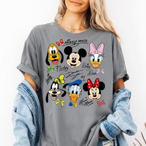 Peut inclure: T-shirt gris avec un motif color&eacute; de Mickey Mouse et ses amis, dont Pluto, Dingo, Donald Duck, Daisy Duck et Minnie Mouse. Le motif comprend les noms et les signatures des personnages. Les manches du t-shirt sont retrouss&eacute;es.