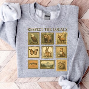May include: A light gray sweatshirt featuring a design of vintage-style postage stamps. The stamps depict various animals and landscapes, with the text "RESPECT THE LOCALS" above the stamps. The stamps include an eagle, bear, deer, butterfly, rabbit, fox, fish, mountains, and squirrel.