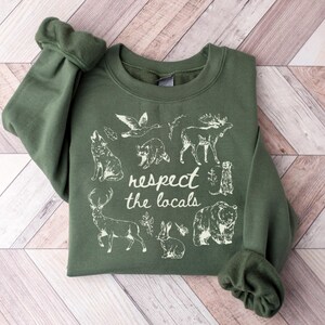 May include: An olive green sweatshirt with the phrase "respect the locals" and illustrations of various animals, including a wolf, bear, and deer. The design is printed in white.