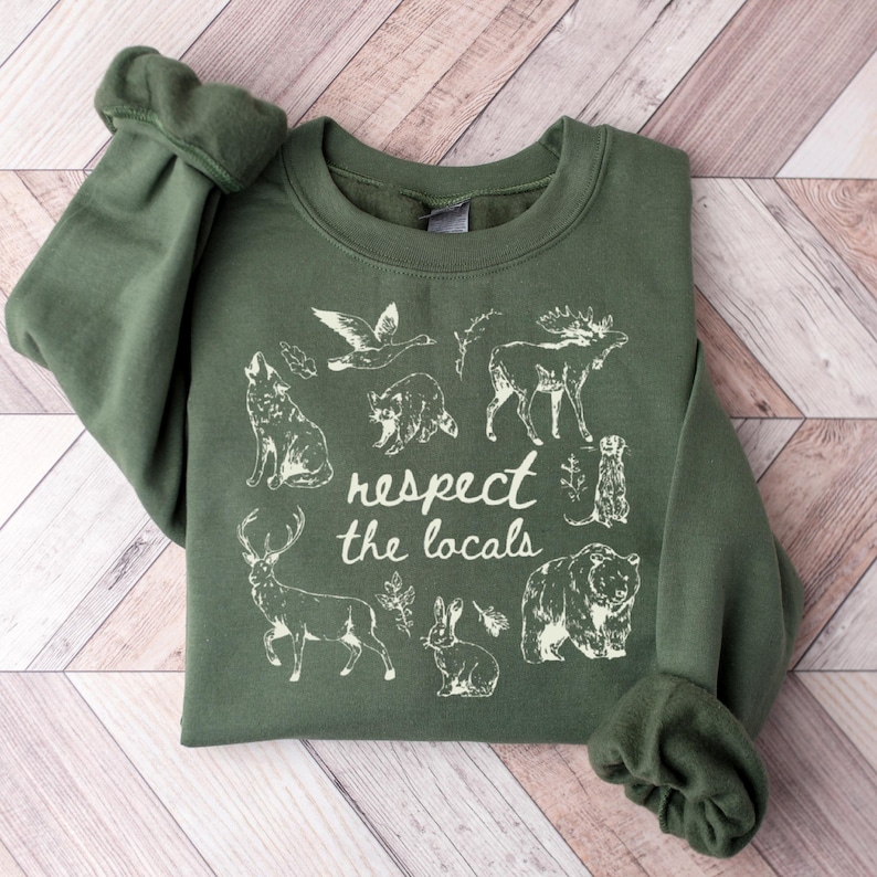 May include: An olive green sweatshirt with the phrase "respect the locals" and illustrations of various animals, including a wolf, bear, and deer. The design is printed in white.